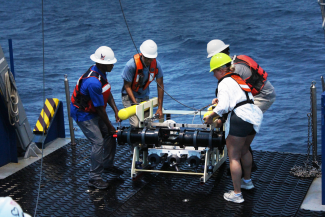 photo off four people with large device in deck of ship, day, water in background