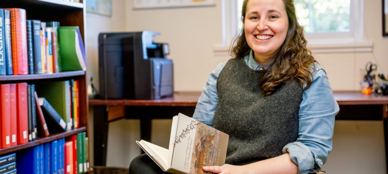 photo of woman sitting in chair with book, books shelves, and window 