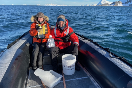 photo of two people in a small boat in water, snow-capped land in background