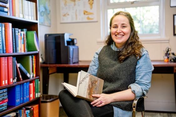 photo of woman sitting in chair with book, books shelves, and window 