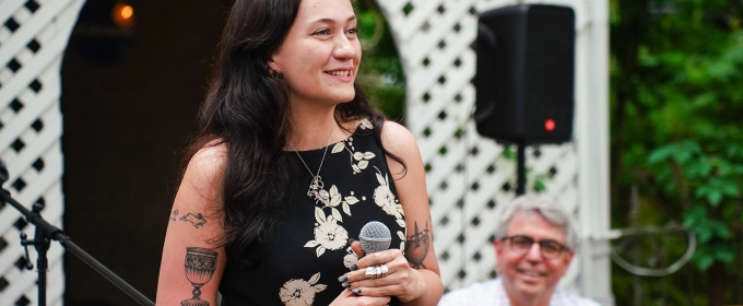 photo of woman with microphone, man in background, outdoors