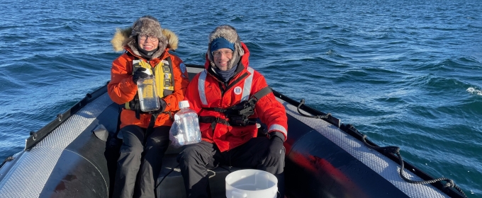 photo of two people in a small boat in water, snow-capped land in background