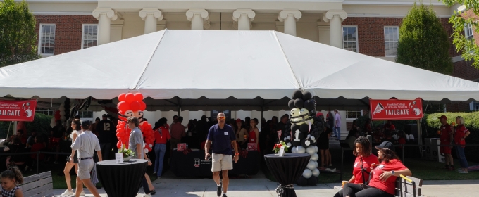 wide photo of tent event with people, building, blue sky