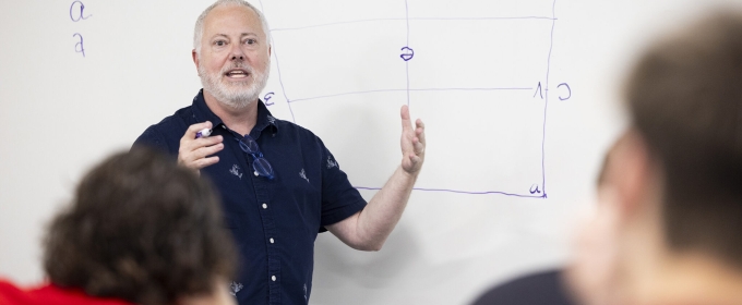 photo of man gesturing, whiteboard in background, students in foreground