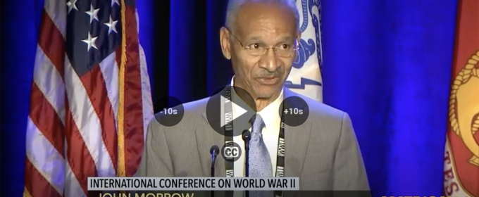 photo of man speaking at microphone, with flags and lower thirds text